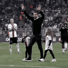 a man stands on a soccer field with his arms in the air while two little girls walk behind him