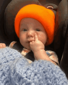 a baby wearing a bright orange hat holds his finger to his nose