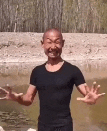 a bald man in a black shirt is standing next to a body of water with his arms outstretched .