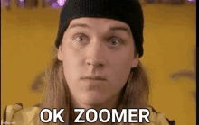 a man wearing a beanie and a yellow jacket is making a funny face and says `` ok zoomer '' .