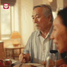 an older man sits at a table with a plate of food and a glass of water with a panda logo in the background