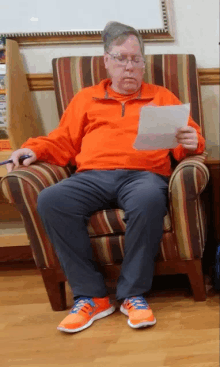 a man in an orange shirt is sitting in a chair reading a letter