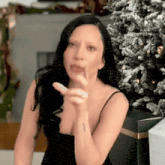 a woman in a black dress is standing in front of a christmas tree and pointing at the camera .