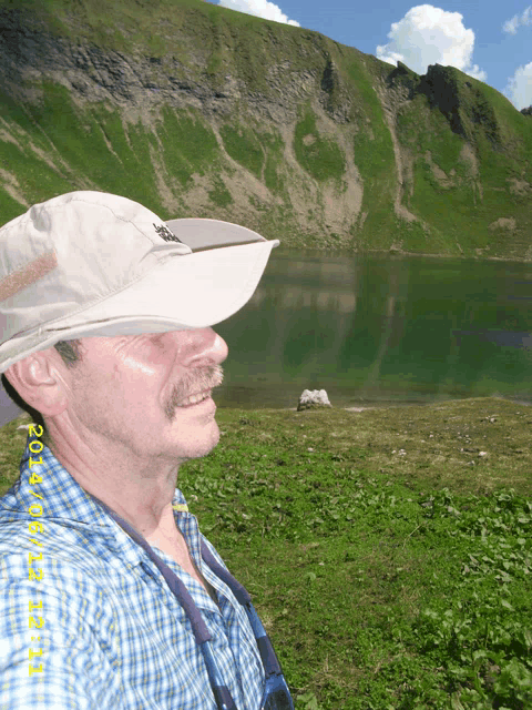 a man wearing a hat stands in front of a mountain and a lake