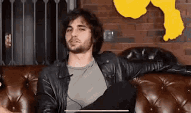 a man in a leather jacket is sitting on a leather couch with his arms crossed .