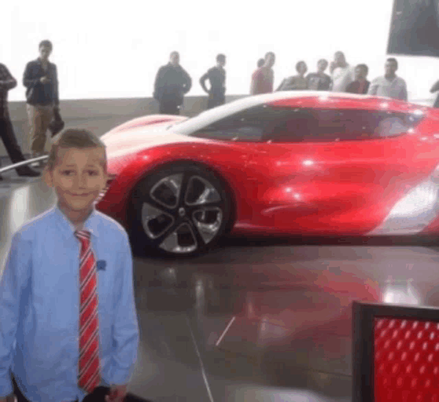 a young boy in a blue shirt and red tie stands in front of a red sports car