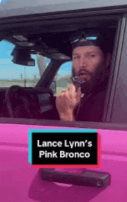 a man is driving a pink bronco and talking on a cell phone .