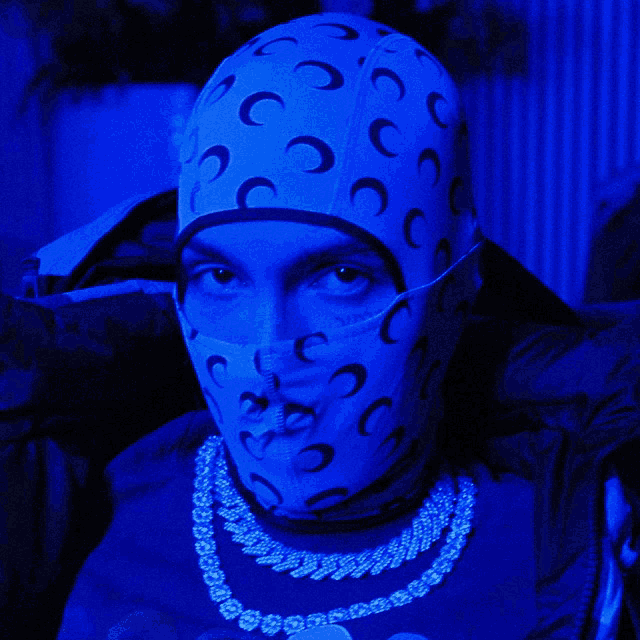 a man wearing a mask and a necklace has a blue background