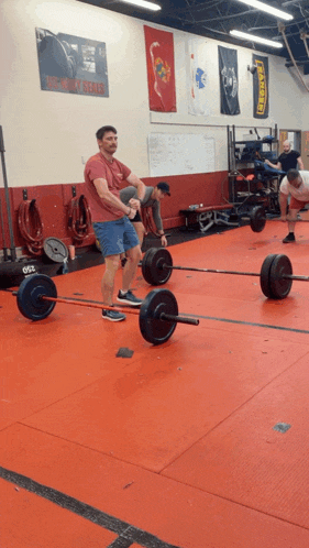 a man in a red shirt is lifting a barbell in a gym with a us navy seals poster on the wall