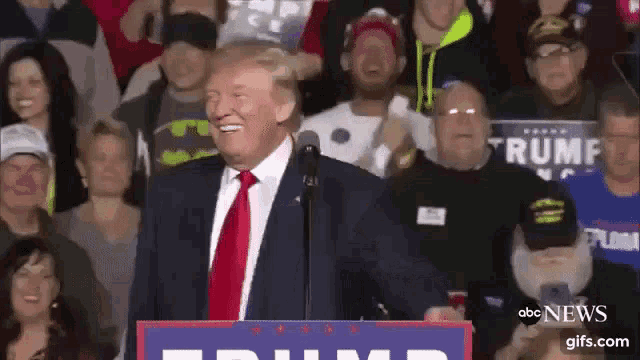 a man in a suit and tie is standing in front of a crowd with a sign that says trump