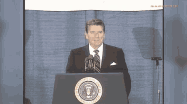 a man in a suit and tie stands at a podium with the seal of the president of the united states on it