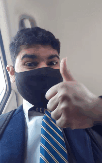 a man wearing a black face mask and a blue and yellow striped tie gives a thumbs up