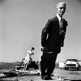 a man in a suit and tie is standing in the dirt in front of a car .