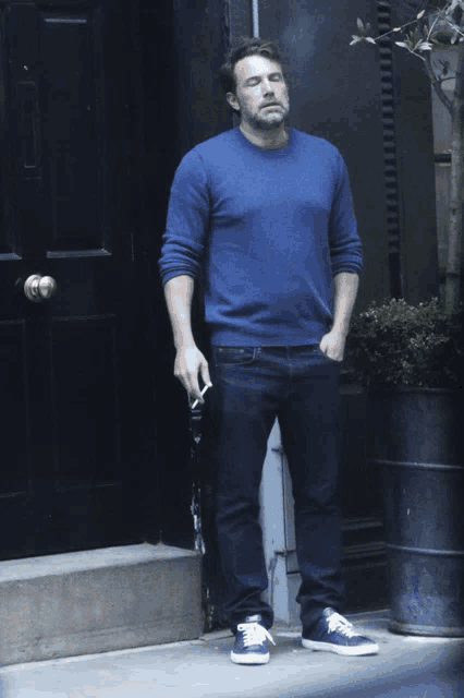 a man in a blue sweater is smoking a cigarette in front of a black door