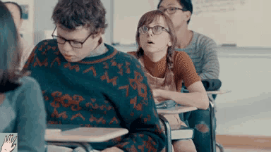 a group of students are sitting at their desks in a classroom and one of them is wearing glasses .