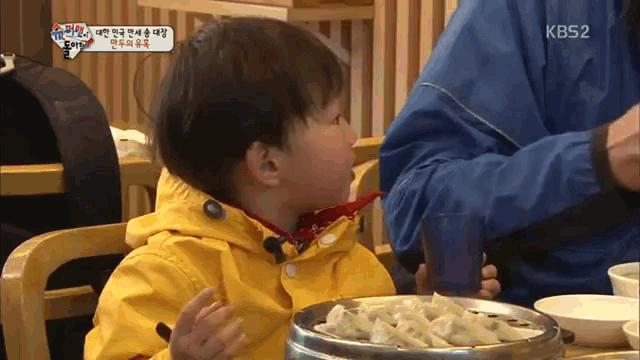 a little boy in a yellow jacket is sitting at a table with a bowl of food and a kb52 logo on the screen