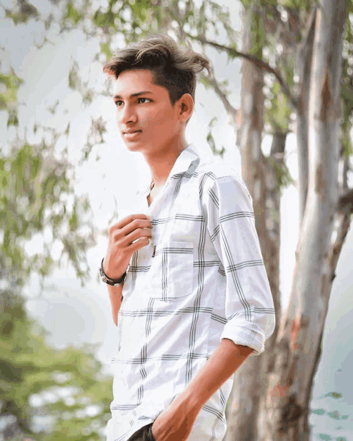 a young man wearing a white plaid shirt stands in front of a tree