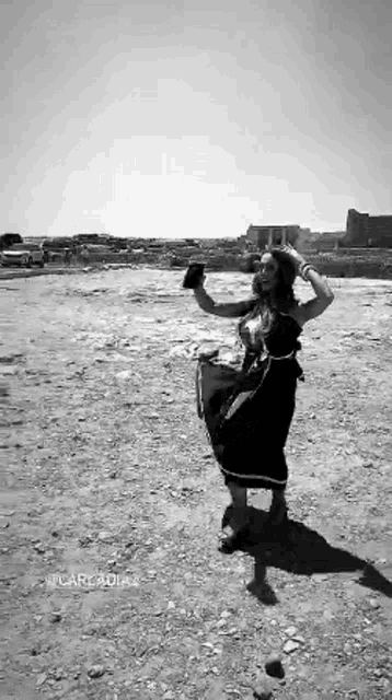 a black and white photo of a woman in a black dress standing in the dirt holding a hat .