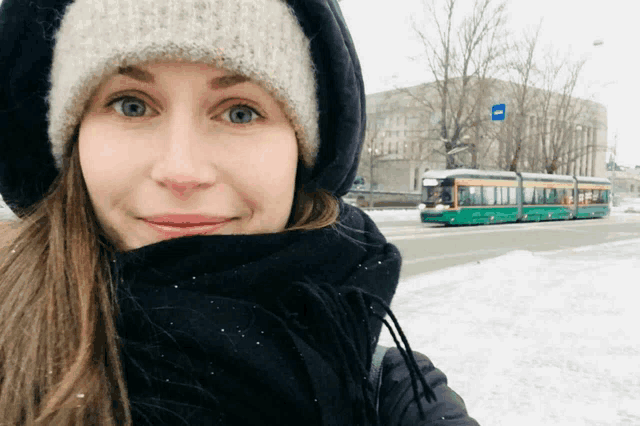 a woman wearing a hat and scarf stands in front of a snowy street with a green bus in the background