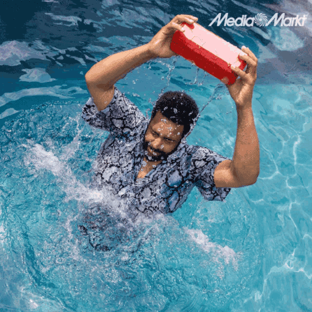 a man in a swimming pool is holding a red box over his head with the media markt logo in the corner