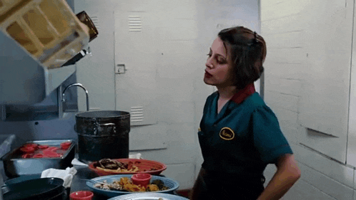 a woman in a green uniform is standing in a kitchen with plates of food and a pot .