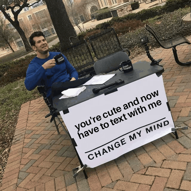 a man in a blue shirt sits at a table with a sign that says change my mind