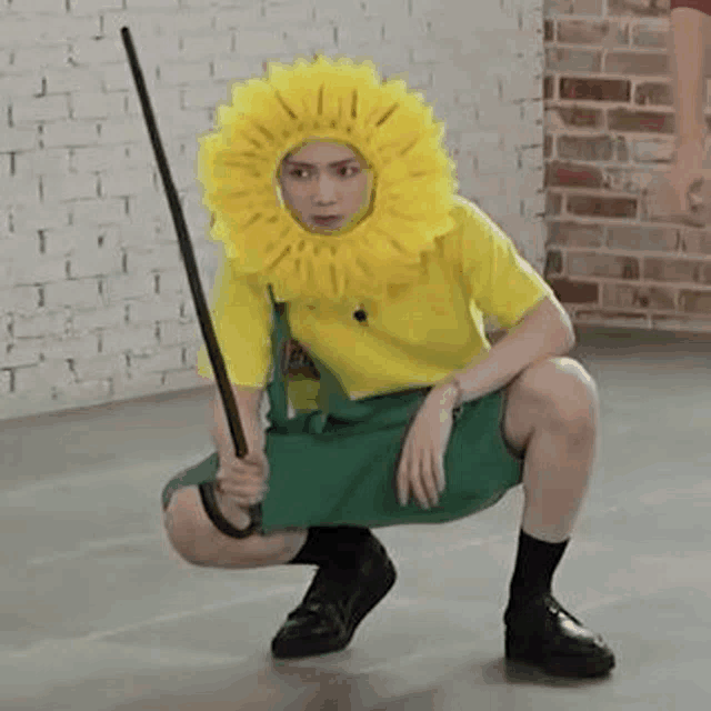 a man wearing a sunflower hat and overalls is squatting down while holding a stick .