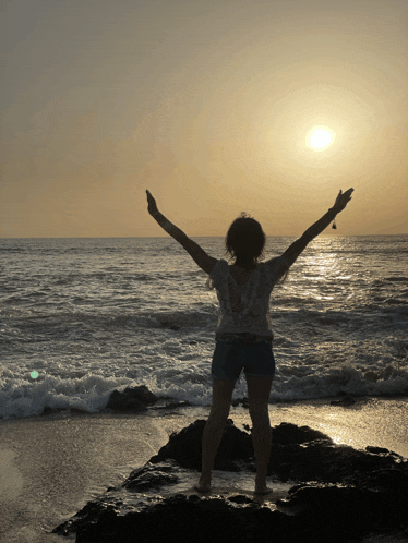 a woman stands on a rocky beach with her arms outstretched at sunset