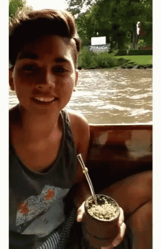 a young man holding a cup with a straw in front of a river with a sign that says itaúda in the background