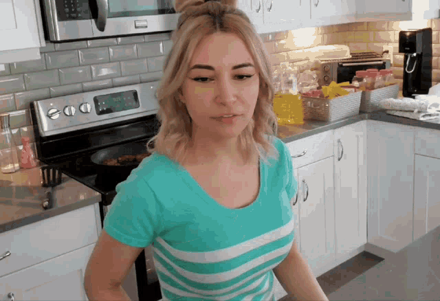 a woman in a blue striped shirt is standing in a kitchen with a stove that has the time of 3:06