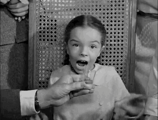 a little girl is sitting in a chair with her mouth open while a man holds a glass of water .