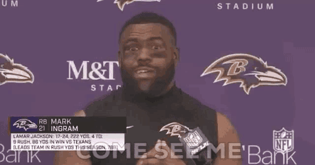 a man with a beard is standing in front of a purple wall with a ravens logo on it