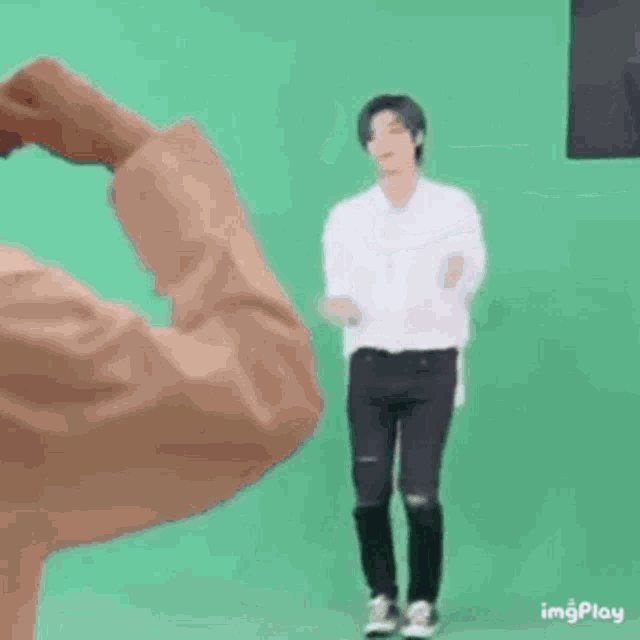 a man in a white shirt and black pants is standing in front of a green screen and making a heart shape with his hands .