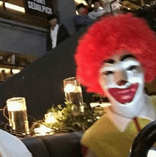 a clown with a red wig is sitting at a table