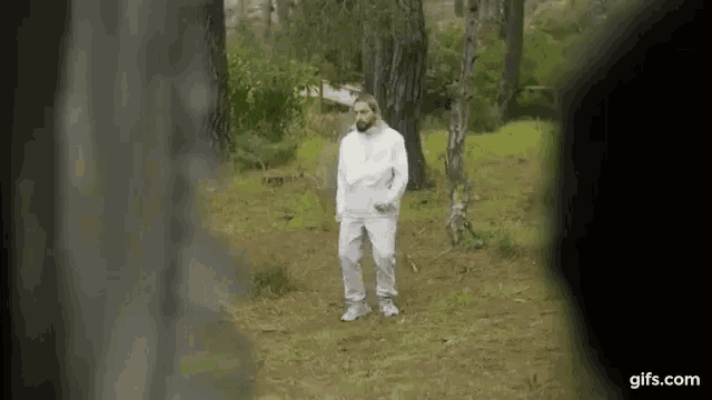 a man in a white hoodie and white pants is standing in a forest .