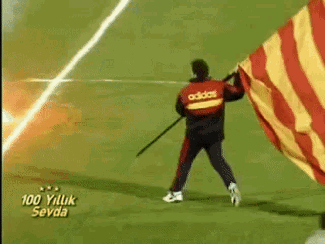 a man in an adidas jacket is holding a flag on a soccer field with smoke coming out of it .