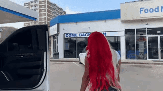 a woman with red hair is standing in front of a gas station with her car door open .