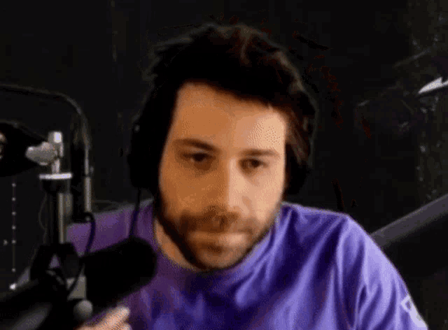 a man with a beard is wearing headphones and a purple shirt while sitting in front of a microphone .