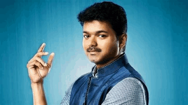 a man with a mustache is standing in front of a blue background and making a hand gesture .