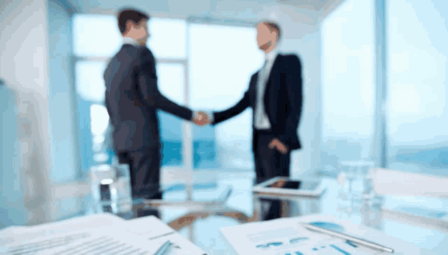 two men shake hands in front of a table with papers and a pen