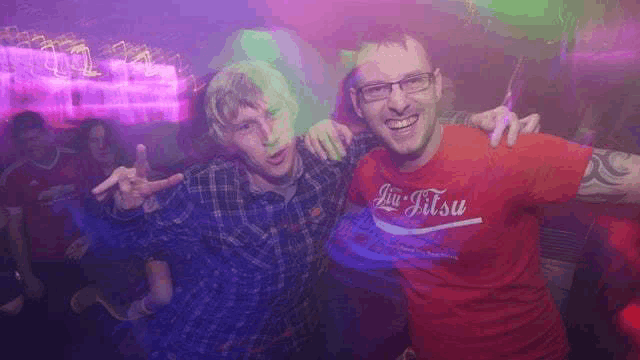 two men are posing for a picture together in a club . one of the men is wearing a jiu-jitsu shirt .