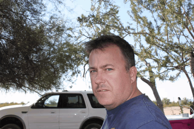 a man in a blue shirt stands in front of a white suv and a tree