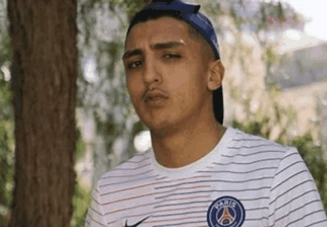 a young man wearing a striped shirt and a blue hat is standing next to a tree .