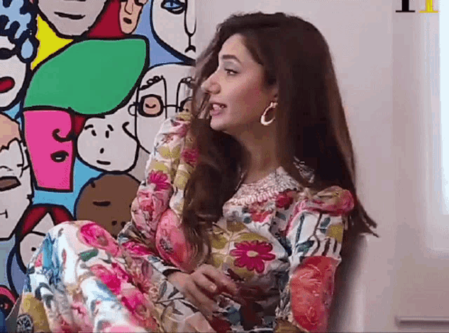 a woman in a floral dress is sitting in front of a wall with drawings of faces on it