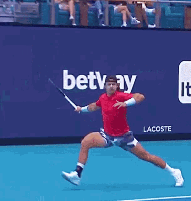 a man in a red shirt is swinging a tennis racquet on a tennis court in front of a lacoste ad