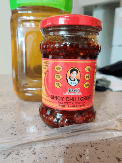 a jar of spicy chili crisp sits on a table next to a bottle of oil