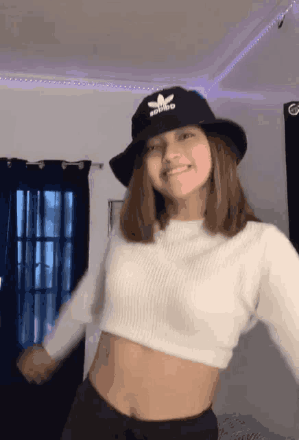 a woman wearing a black adidas hat and a white crop top is dancing in a room .