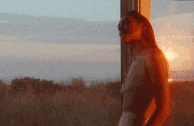 a woman stands in front of a window looking out at the sunset