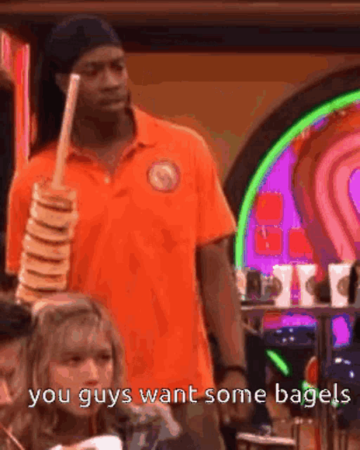 a man in an orange shirt is holding a stack of bagels and says " you guys want some bagels " .