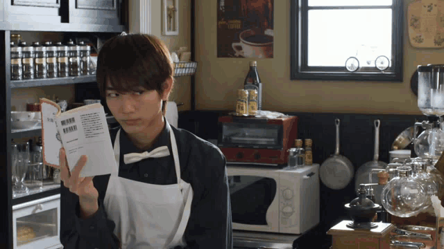 a man in an apron is reading a book in a kitchen with a coffee poster on the wall behind him